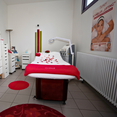 Behandlungsraum mit Liege mit rotem Bezug und Kosmetikgeräten.